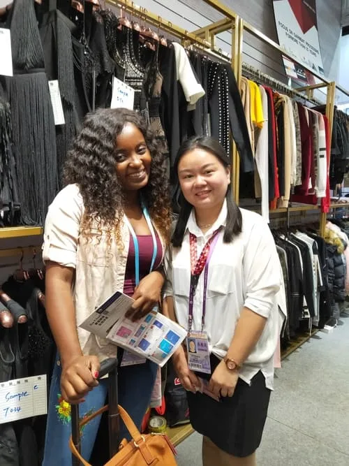 Two women smiling in a clothing store, standing before racks of garments reminiscent of a Canton Fair display. One holds papers and wears a lanyard, while the neatly hung autumn clothing in the background adds an exhibition-like charm.