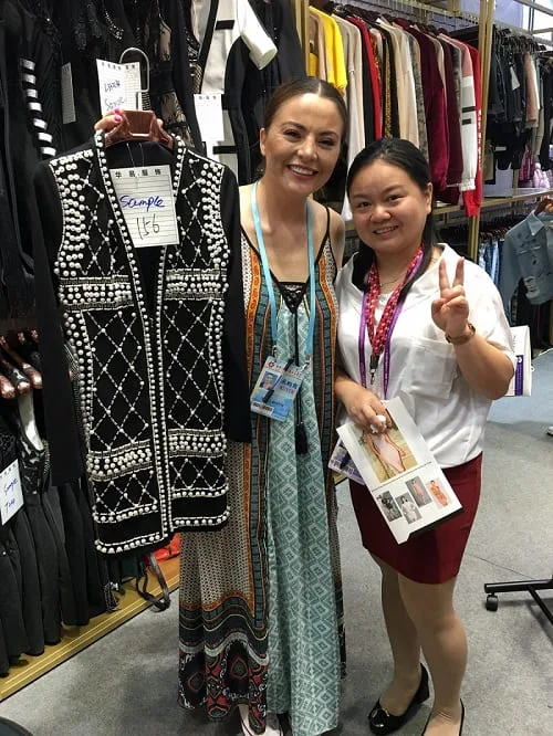 Two women smile in a clothing store, reminiscent of the vibrant scenes at the Canton Fair. One holds a sleeveless black and white dress labeled Sample $46, while the other flashes a peace sign with images of clothing, surrounded by racks of stylish garments.