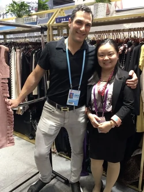 A man and a woman stand smiling in front of a clothing rack displaying various garments at the Canton Fair. The man wears a black polo shirt and light pants; the woman is dressed in a black blazer and black dress with a pink necklace, capturing the essence of this prominent clothing fair in May.