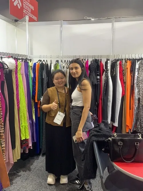 Two people stand in a clothing store, smiling. One is giving a thumbs-up while holding a name tag, reminiscent of those at the Melbourne Clothing Trade Show. Various colorful dresses hang on racks in the background. A black handbag sits on a chair in the foreground.
