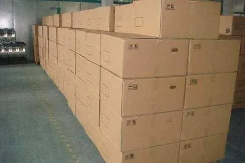 Stacks of large cardboard boxes are neatly arranged in rows inside a warehouse. The floor is smooth concrete, and the boxes are uniform in size and color, with some markings on their sides.