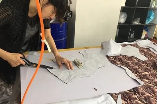 Person using scissors to cut fabric patterns on a table, with patterned and plain fabric pieces. An orange hose hangs nearby, and shelves with various items are in the background.