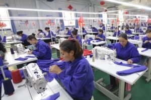 People in blue uniforms work at sewing machines in a well-lit factory, crafting purple fabric for the bandage dress production line. The room is organized with rows of workstations, and small red flags adorn some tables.