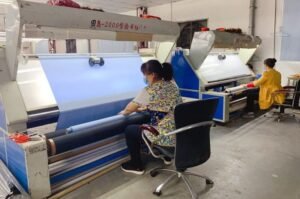 Two people seated at separate fabric inspection machines in a factory setting. They are examining large rolls of blue material under bright lights. The floor is concrete, and theres a metal chair nearby.