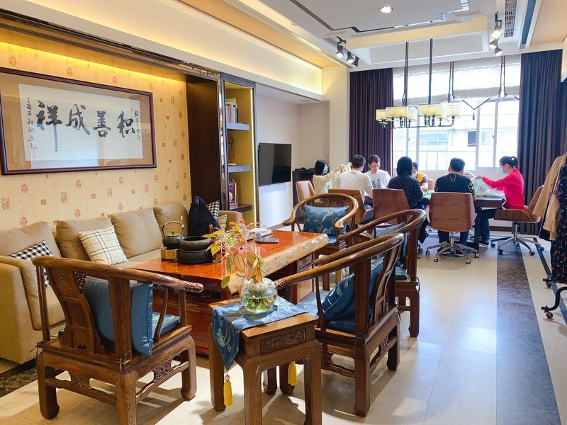 A cozy living and dining area with traditional wooden furniture. A calligraphy artwork adorns the wall above a sofa. A group of people is seated around a table by large windows, engaged in conversation. Bright, natural light fills the space.