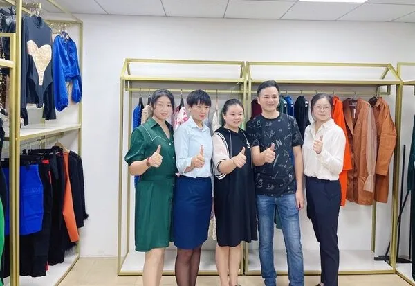 Five foreign trade elite team members stand in a clothing store, giving thumbs up. The background showcases racks of colorful garments. Their smiles create a positive and cheerful atmosphere, highlighting their success and camaraderie.
